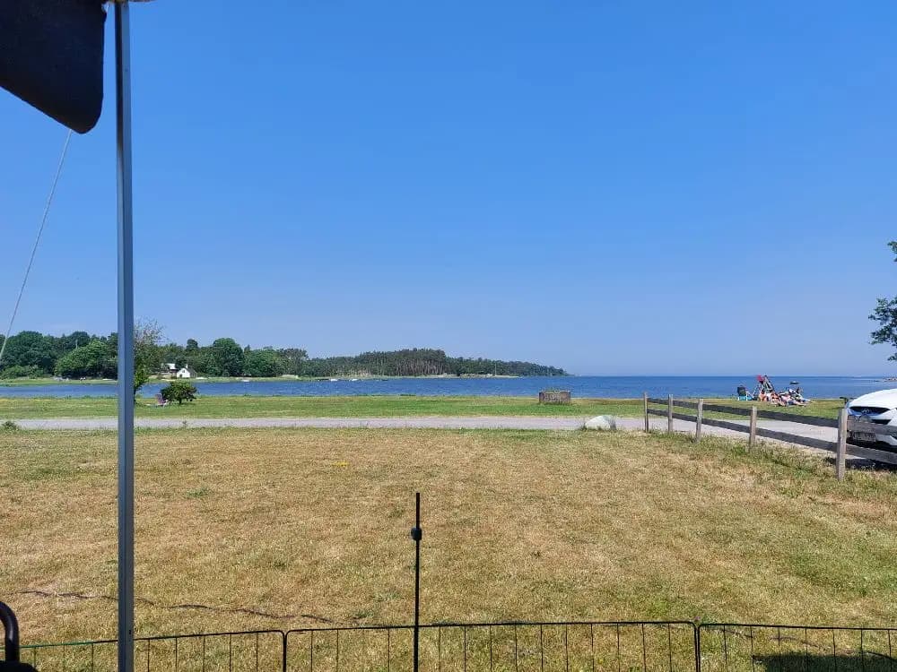 Bild av Skeppeviks camping: En vidsträckt gräsyta med skugga under träd, ett staket som sträcker sig längs kusten, och parkerade husvagnar syns i bakgrunden. Himlen är klarblå.