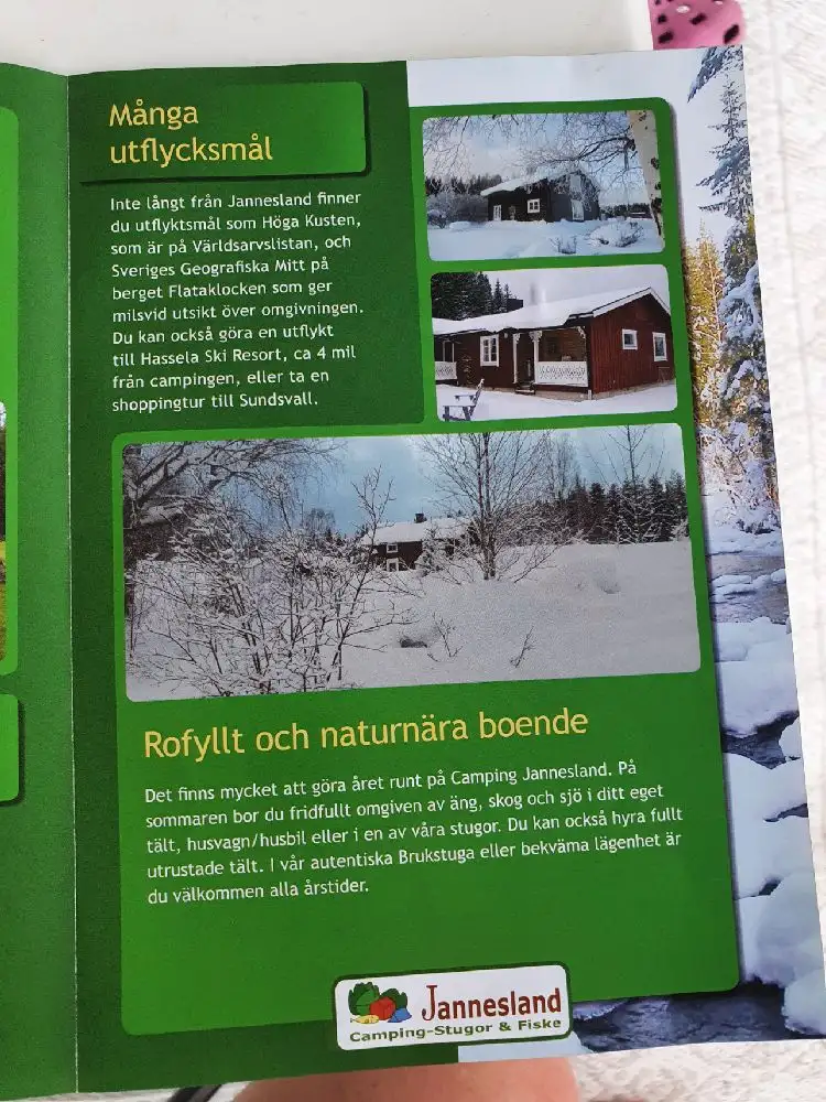 En snötäckt stuga på campingplatsen Jannesland, omgiven av vinterlandskap. Stugan är byggd i traditionell stil med timmerstockar, och en liten vedbod syns bredvid. Marken är täckt av ett tjockt lager snö, vilket förstärker den fridfulla vinterkänslan.