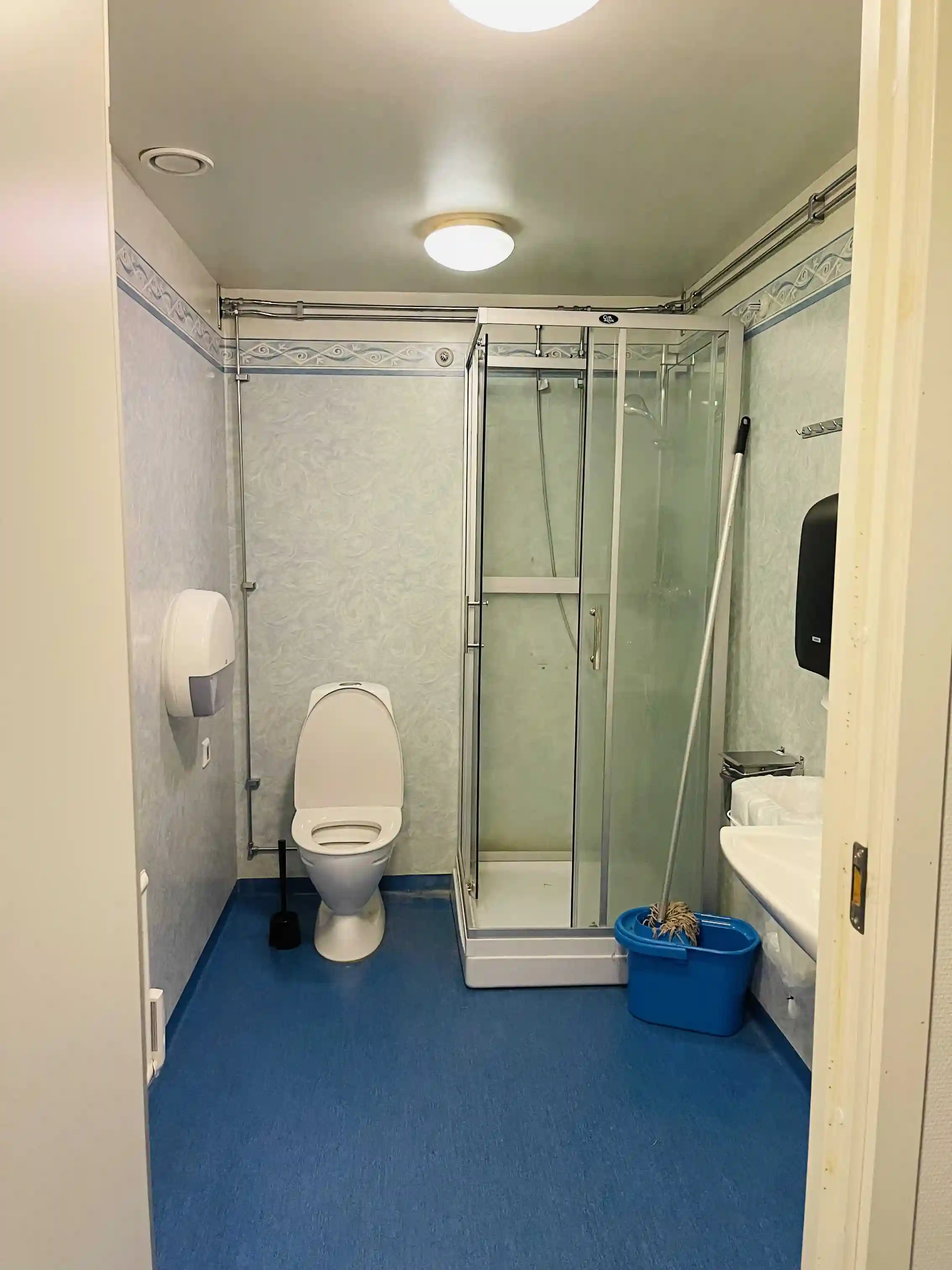 En modern och ren badrumsinteriör på Hjälmaresunds camping, med ett ljust kaklat golv och väggar. Ett vitt handfat med blandare och en spegel finns på ena sidan, och en vit toalettstol är placerad bredvid. En dörr syns i bakgrunden, och ett vitt tak kompletterar det ljusa rummet.