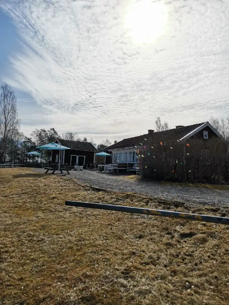 En lantlig campingplats vid Hjälmaresund med en stuga i förgrunden, omgiven av öppna landskap och en solbelyst himmel med moln.