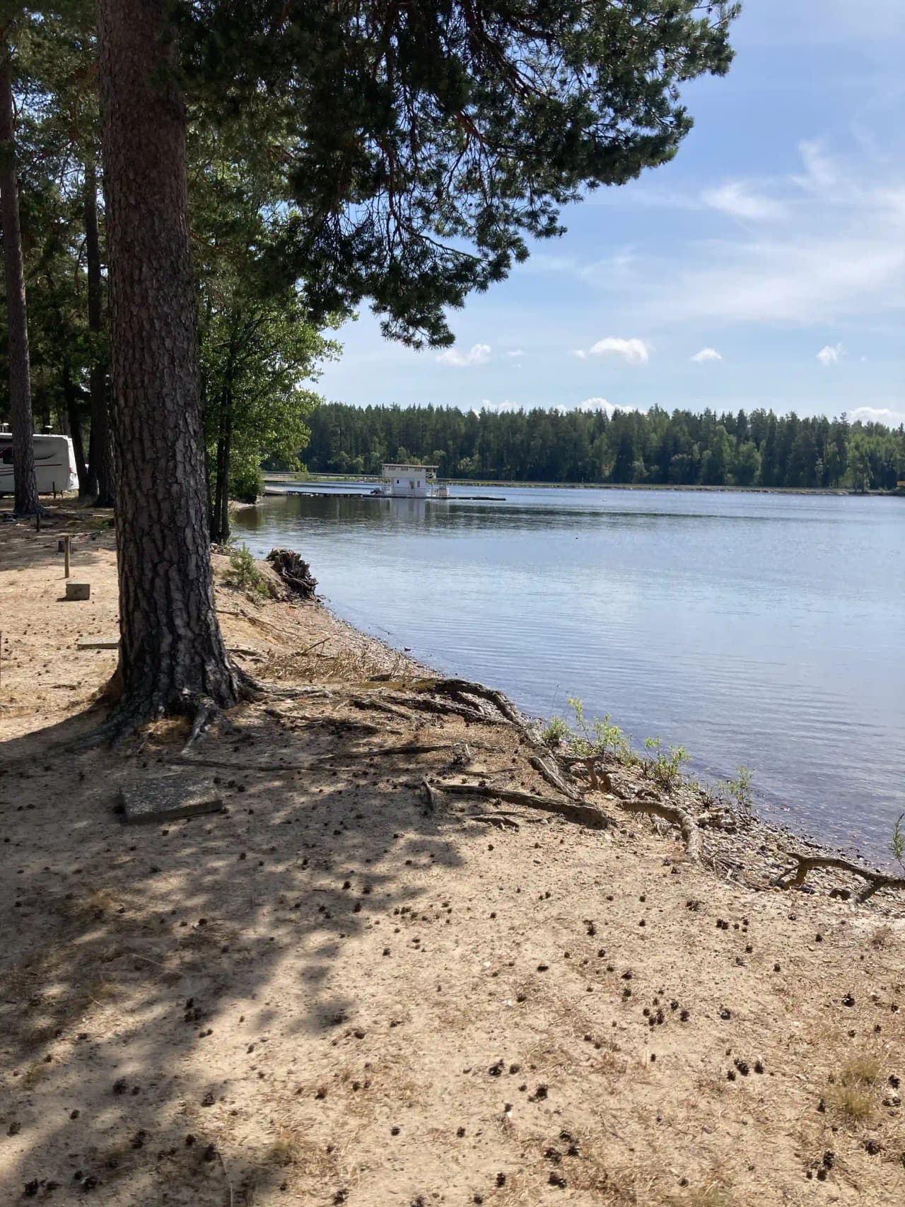 En vy av Revelbadets camping som visar en sjö med en strandlinje. I bakgrunden syns träd och en klar himmel, vilket skapar en naturskön och lugn atmosfär.