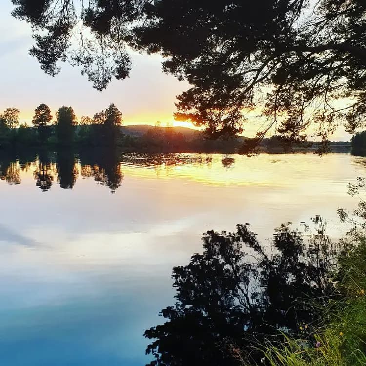 En stilla vattenyta vid Nås camping, omgiven av träd och natur. Skymningen sänker sig över landskapet och speglas i vattnet.