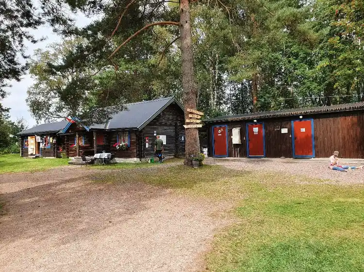 En grönskande campingplats med träd, skuggiga områden och små stugor. En loge och andra trädgårdsbyggnader syns i bakgrunden i en park-liknande miljö.