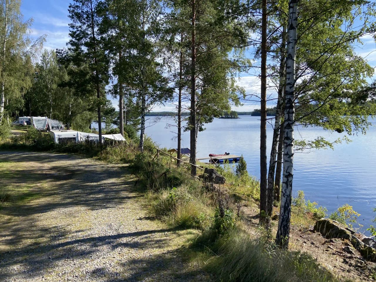 Bild av Vammervikens camping: En vacker sjö omgiven av frodig grönska och höga träd, vilket skapar en naturskön och lugn miljö för avkoppling och friluftsliv.
