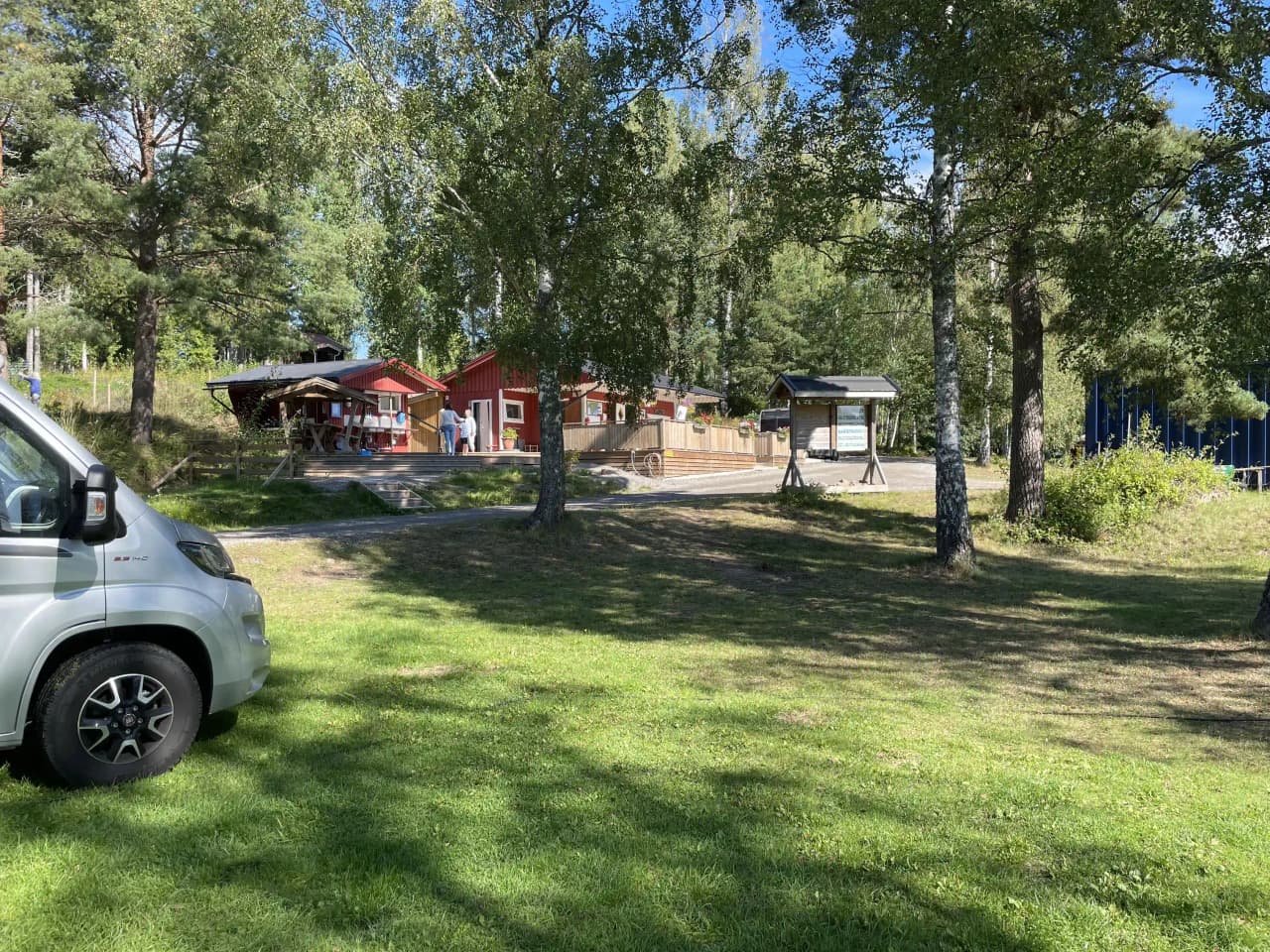 En grönskande campingplats med träd och växter i bakgrunden. Skuggiga områden ger avkopplande miljö, och en stor gräsmatta syns med parkeringsområde intill. Trädgårdsbyggnader ligger utspridda på området.