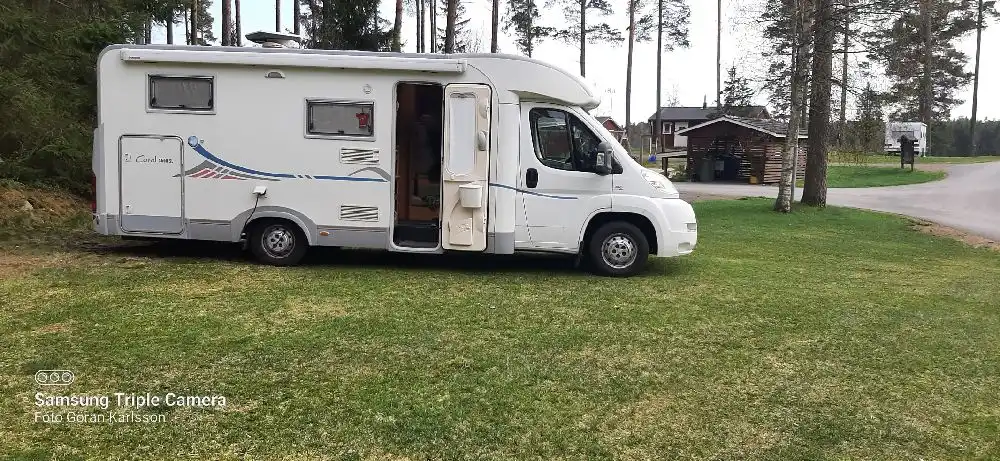 En husbil parkerad vid Hoks naturcamping, omgiven av naturskön miljö. Bilen är försedd med sidobackspeglar, registreringsskylt och andra vanliga fordonsdetaljer som kaross och stötfångare.
