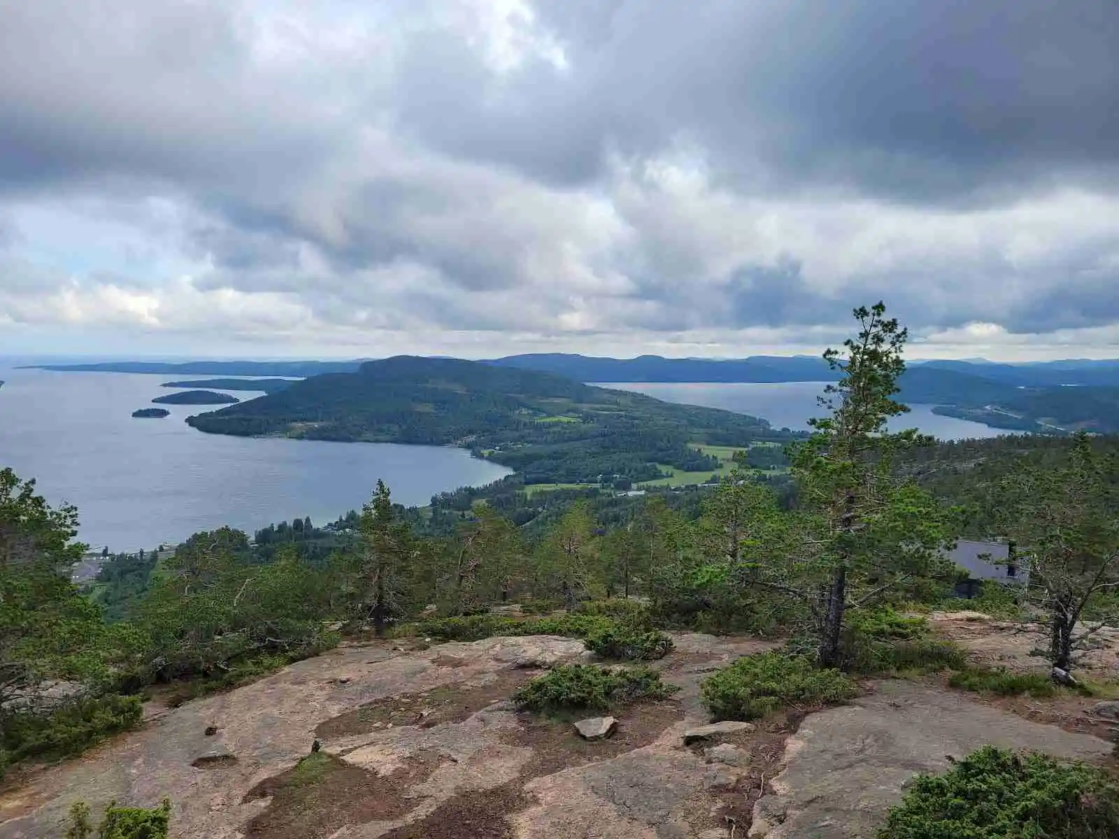 Bild av en naturskön plats vid Docksta vandrarhem & camping Kustladan, med en vidsträckt vy över ett kuperat landskap. I förgrunden syns en sjö och kustlinje, omgiven av gröna höjder och skog. Moln täcker delvis en blå himmel och förstärker den vilda naturens inbjudande atmosfär.