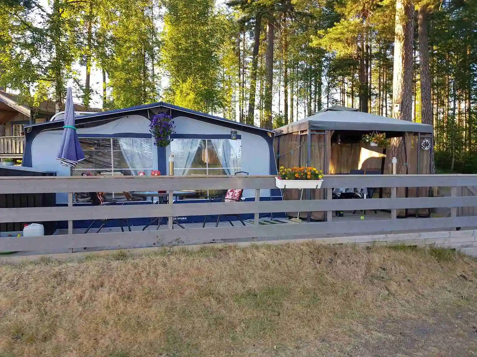 Bilden visar en naturskön campingplats vid namn Treens Natur & Fiskecamp. Scenen bjuder på skuggiga ytor med träd som omger platsen, och i bakgrunden syns enkla trädgårdsstrukturer samt mobila hem. En lugn och fridfull atmosfär i en välordnad bakgård med en inbjudande, naturnära miljö.