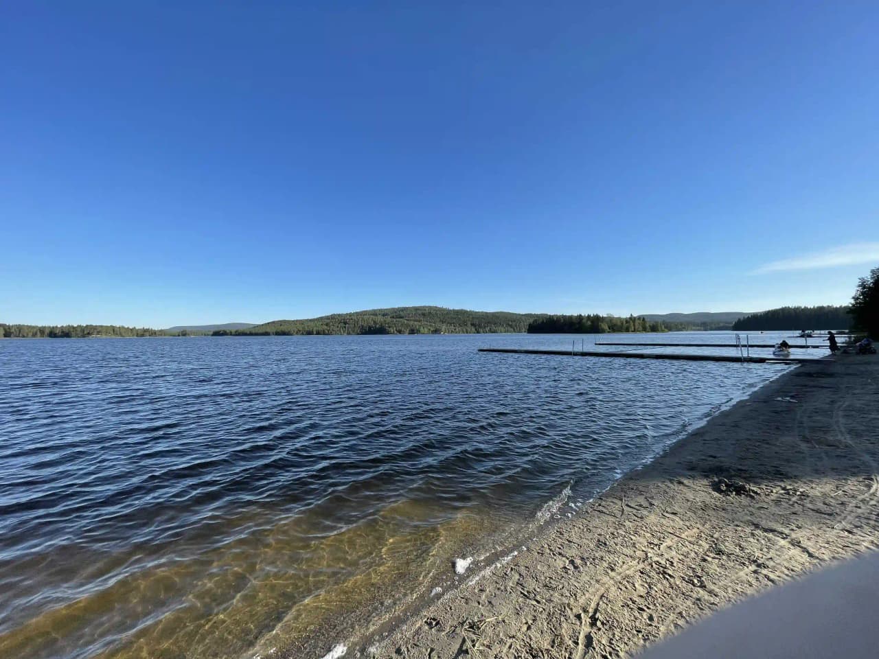 En idyllisk vy över Storsjö camping & trädgård med en klarblå himmel som speglas i det stilla vattnet, horisontlinjen sträcker sig bortom sjöns kustlinje.