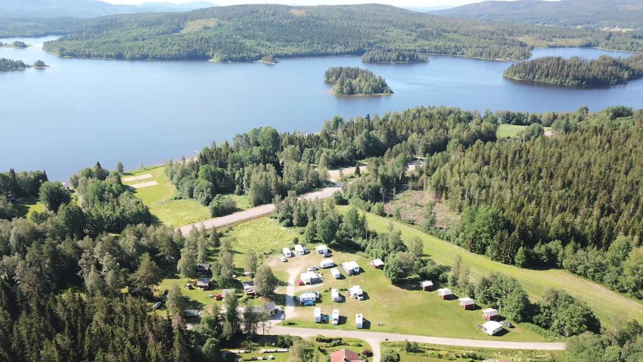 Flygbild över Storsjö Camping & Trädgård, visar en naturskön miljö med träd, växtlighet och en sjö. Vattendrag rinner genom området omgivet av skog och landskap.