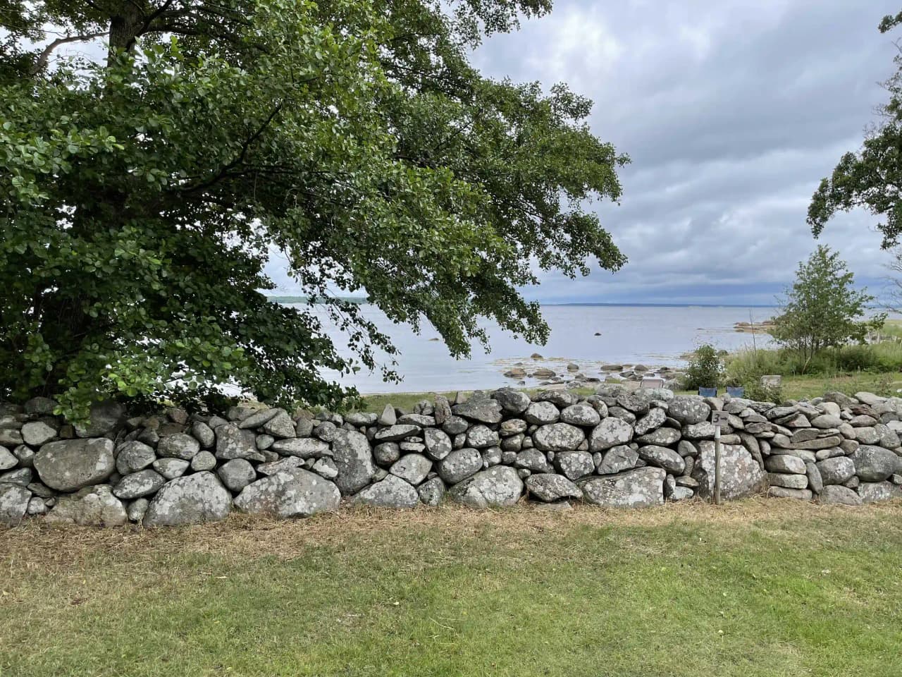En stenmur vid en kustlinje, omgiven av grönt gräs och naturligt landskap. En vacker utsikt över en stenig strand och kustens oregelbundna formationer kan ses i bakgrunden.