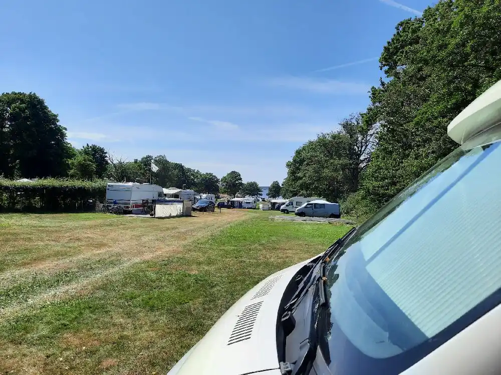 En bild av en parkeringsplats vid Stenbräckagården Hägnan, med flera fordon som bilar, husbilar och en van. Fordonen är uppställda på platsen, omgivna av träd och natur.