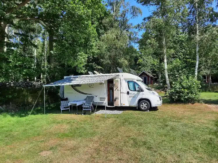 En campingplats med flera husvagnar och tält uppställda i friluftsmiljö vid Stenbräckagården Hägnan, omgiven av en parkering med bilar och en grönskande gård.