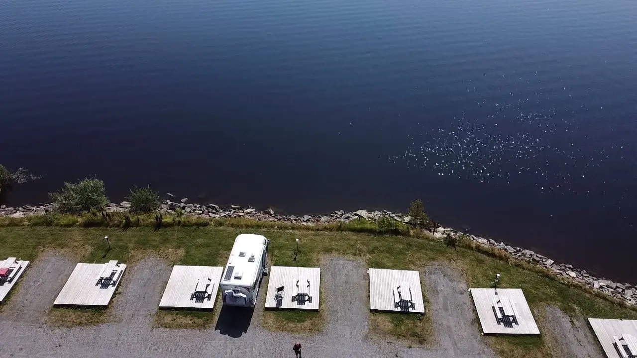 Flygbild över Arvika småbåtshamn med flera husbilar parkerade vid vattnet. Vattnet är kantat av grönska och skapar en rofylld atmosfär som påminner om en sjödistrikt.