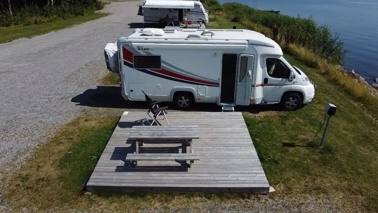 En campingplats vid Arvika småbåtshamn med flera husbilar och husvagnar parkerade intill varandra, omgivna av grönska och vatten i bakgrunden.