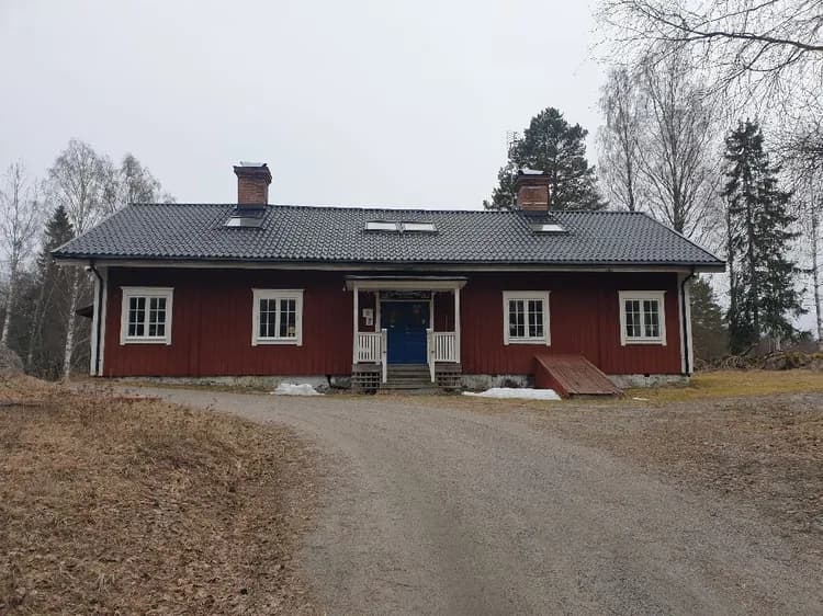 En röd stuga med vitmålade fönster och dörrar, belägen på en lummig tomt i ett lantligt område. Stugan har ett sluttande tak och omgivningen ger en känsla av landsbygd och natur.
