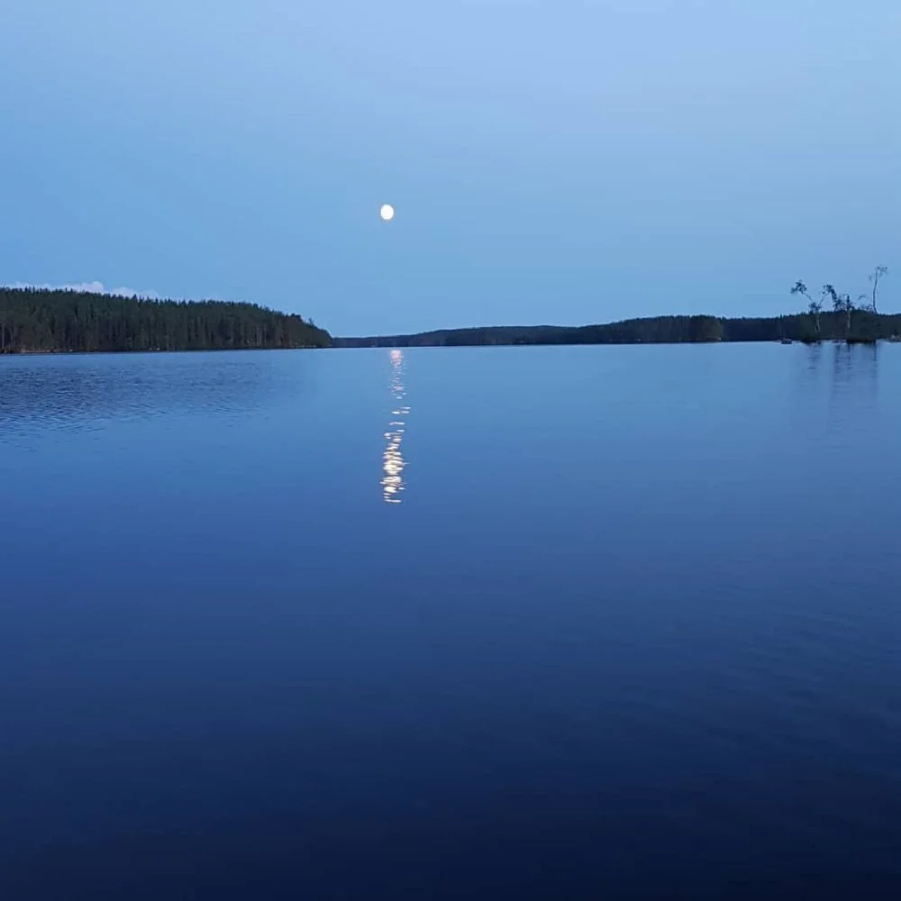 En stilla sjö med blått vatten där månen reflekteras på ytan, omgiven av en horisont i fjärran.
