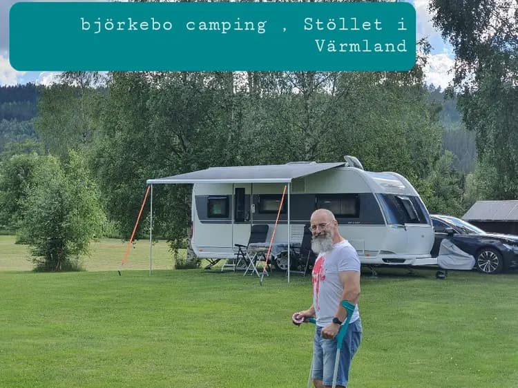 Bild av Björkebo Camping med husbilar uppställda på en grön, solbelyst äng omgiven av träd. En semi-trailer truck syns i bakgrunden, och en gräsmatta med tält ligger i närheten av en liten park.