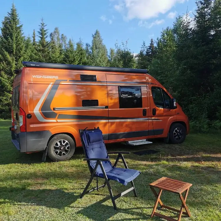 En vit husbil med bilens sidodörr öppen, parkerad på Svennevads camping. Husbilen är utrustad med en bagageräcke på taket och står intill en grönskande miljö.