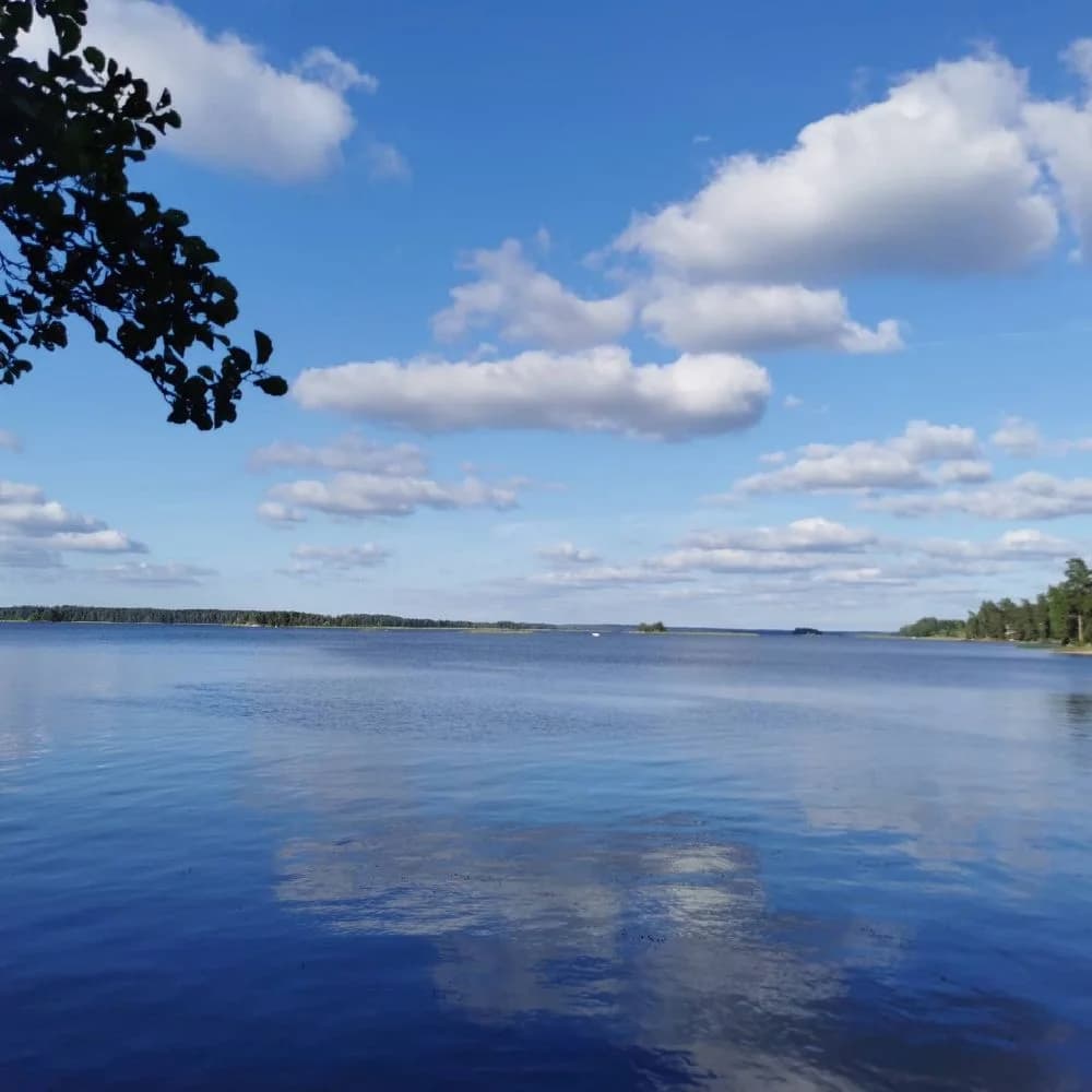 En vidsträckt blå himmel med vita moln speglas i en stilla vattenyta vid Svennevads camping. Horizonten skymtas i fjärran under en klar dag.