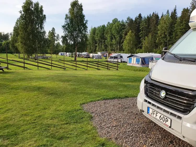 En husbil och en husvagn står parkerade på en grön gräsmatta vid Svennevads camping, omgivna av träd och en liten parkering.