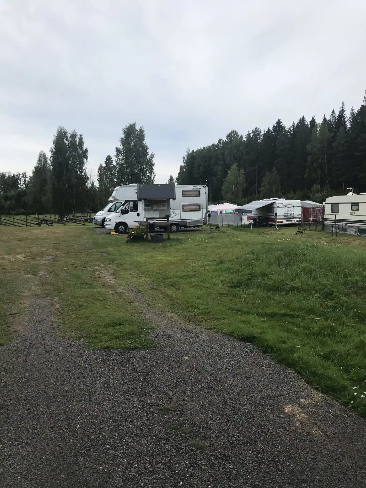 En parkeringsplats på Svennevads camping fylld med husbilar och bilar, inklusive en husvagn, omgiven av träd och grönska.