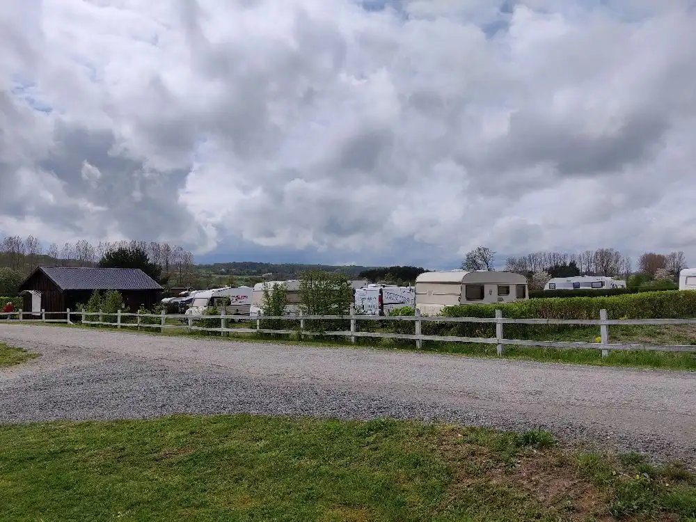 En vidsträckt äng under en klar himmel med vita moln, omgiven av ett trästaket som leder till en campingplats.