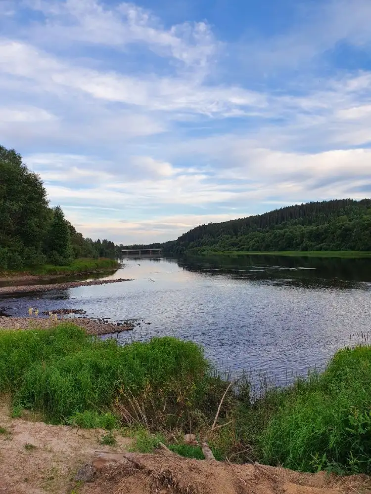 Vy över Värnäs camping med en vacker naturmiljö, där en flod flyter genom landskapet, omgiven av gröna träd och en klarblå himmel med lätta moln.