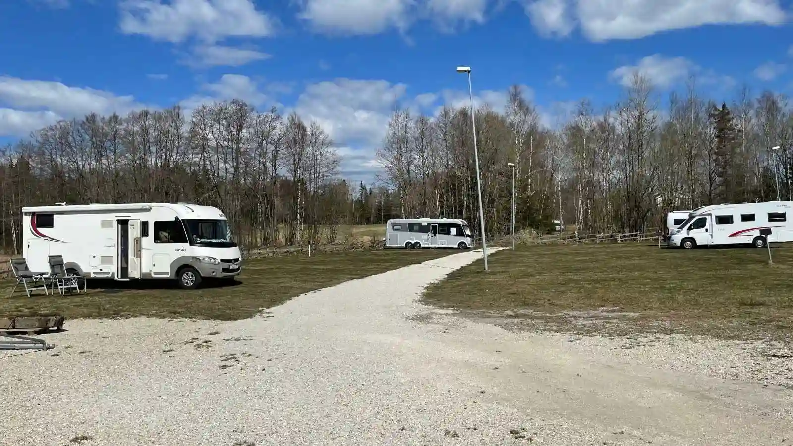 En campingplats med flera husbilar och husvagnar uppställda på rad, omgivna av grönska och parkerade på grusunderlag.