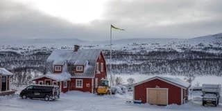 En vinterlandskap vid Fjällnäs Camping, täckt av snö och is, med sluttande terräng och fjäll i bakgrunden under en klar himmel.