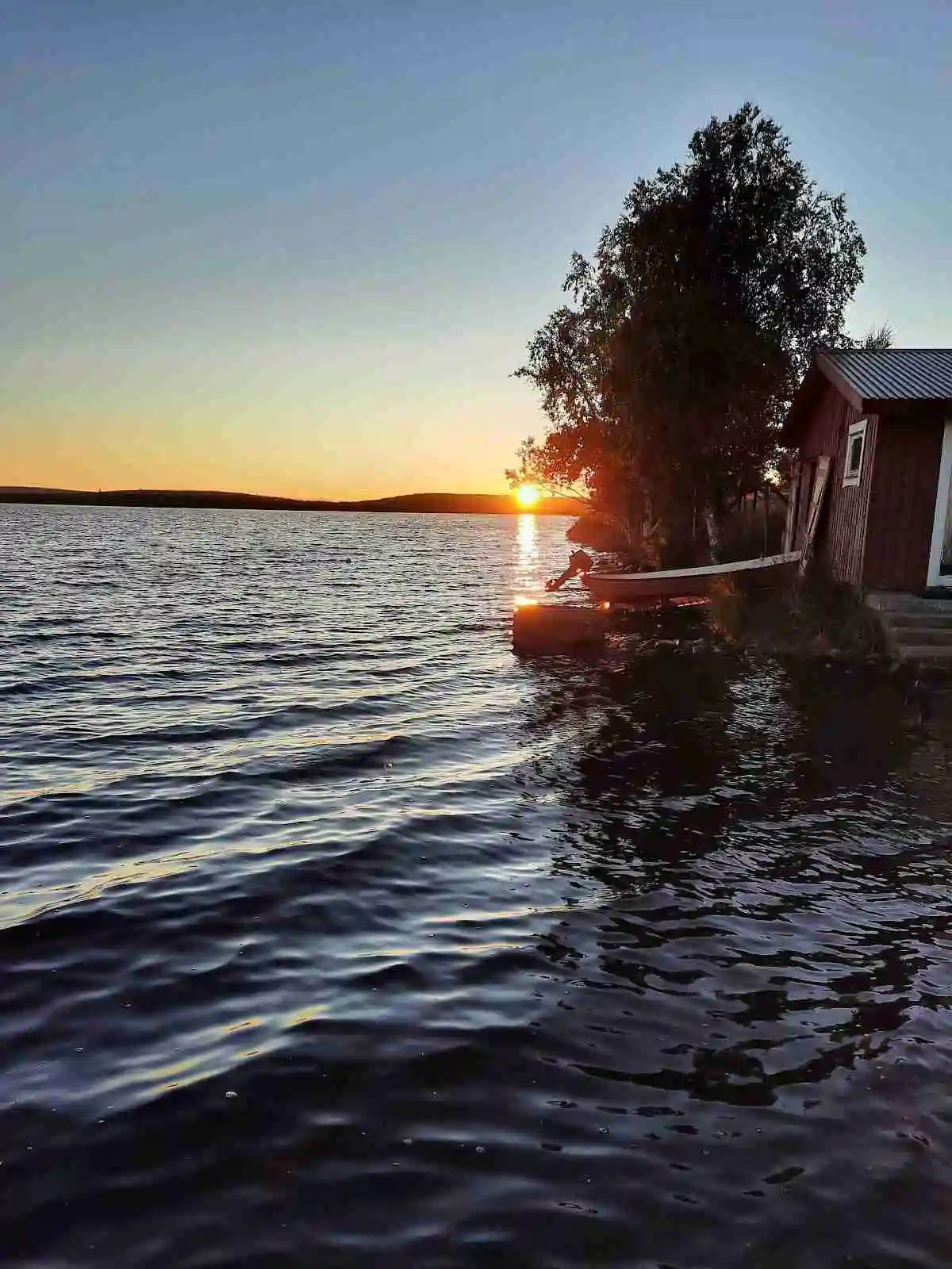 En rofylld scen vid Piilijärvi Camping vid skymningen, med en sjö i förgrunden och en båt som ligger stilla på vattnet. Horisonten badar i mjuka färger från solnedgången, vilket skapar en stämningsfull atmosfär.