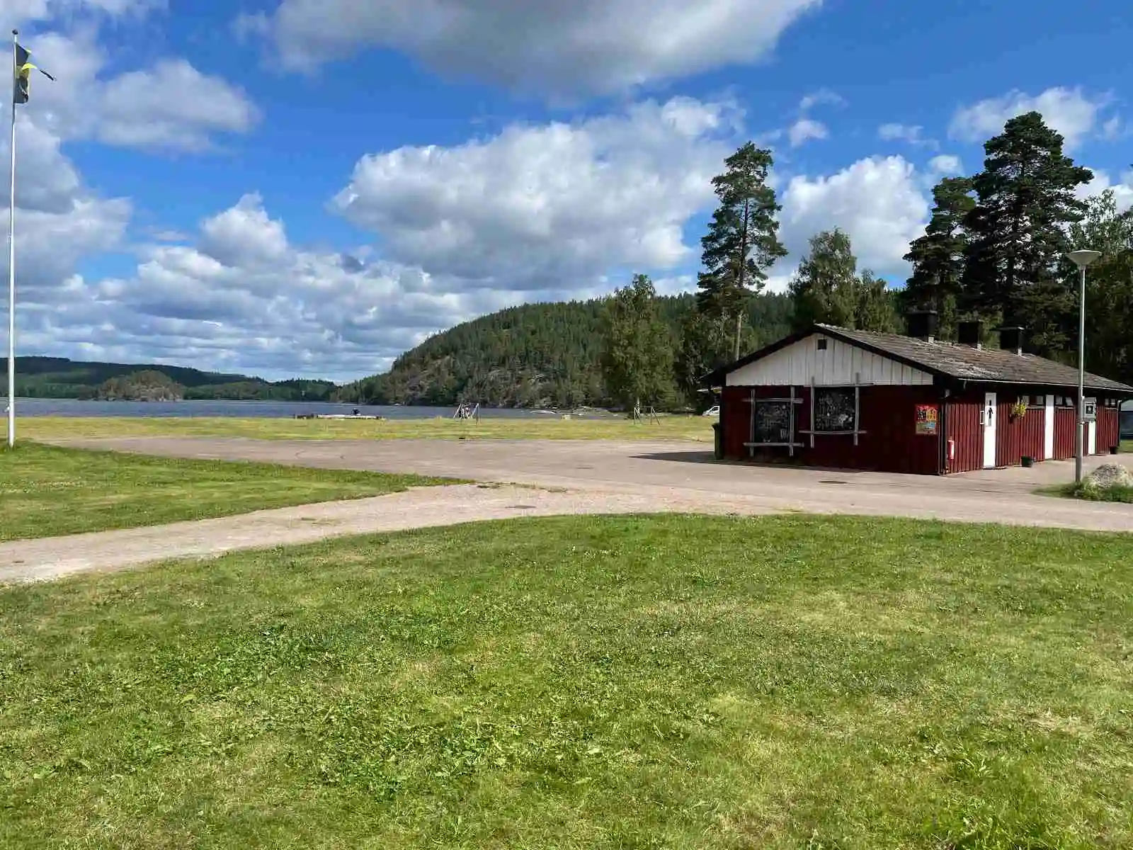 En grönskande campingplats med stora gräsytor och skuggiga områden, som erbjuder plats för tält och husvagnar. I bakgrunden syns stängselstolpar och en anslutande uppfart.