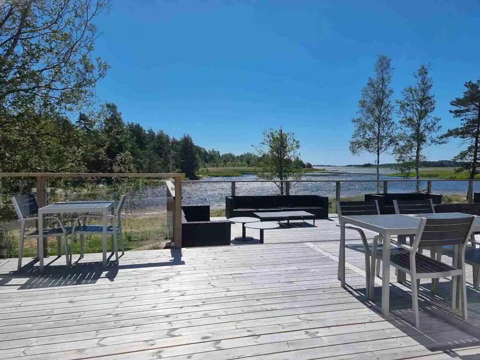 En träbänk och ett bord står på en träplattform vid kanten av en sjö vid Ekenäs gästhamn & camping. I bakgrunden syns det glittrande vattnet, omgiven av gröna träd.