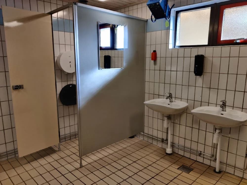 En ljus och modern badrumsmiljö med golvklinker och vita väggar. Till vänster syns en handfat med en silverkran och under handfatet finns förvaringsutrymme. Högersidan domineras av en dusch med glasvägg. Belysningen är diskret och kompletterar den fräscha interiören.