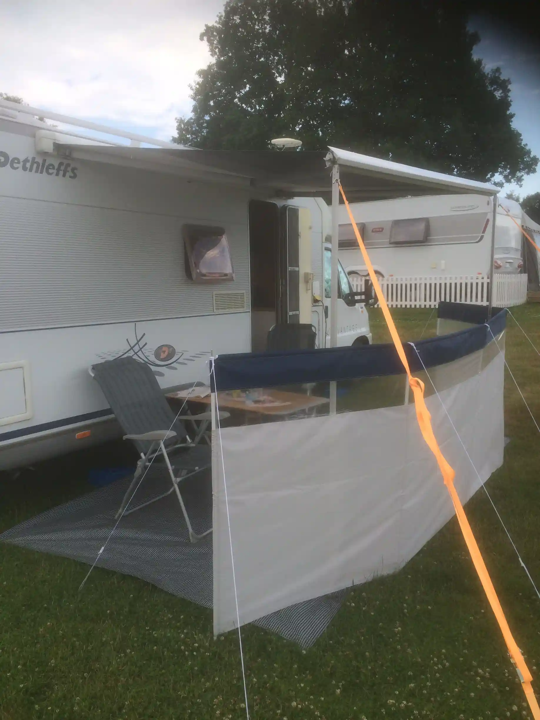 En tältduk hänger över ett skuggigt område vid Järnaviks camping, med en husvagn synlig i bakgrunden. Det blåser lätt i vinden.