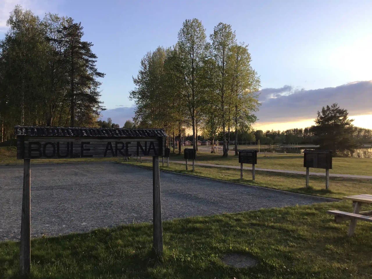 En grönskande campingplats i Storuman med träd och gräsytor. Morgonsol skiner genom trädkronorna, och en parkbänk står i skuggan.