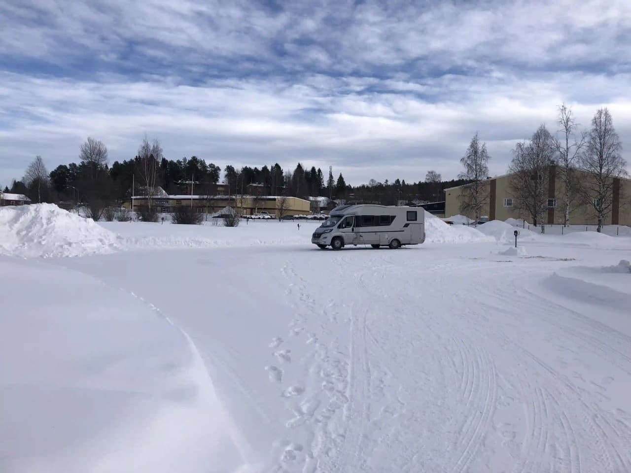 Snötäckt campingområde med parkerade bilar vid skogskant. Stora snötäcken, vinterlandskap med barrträd i bakgrunden.