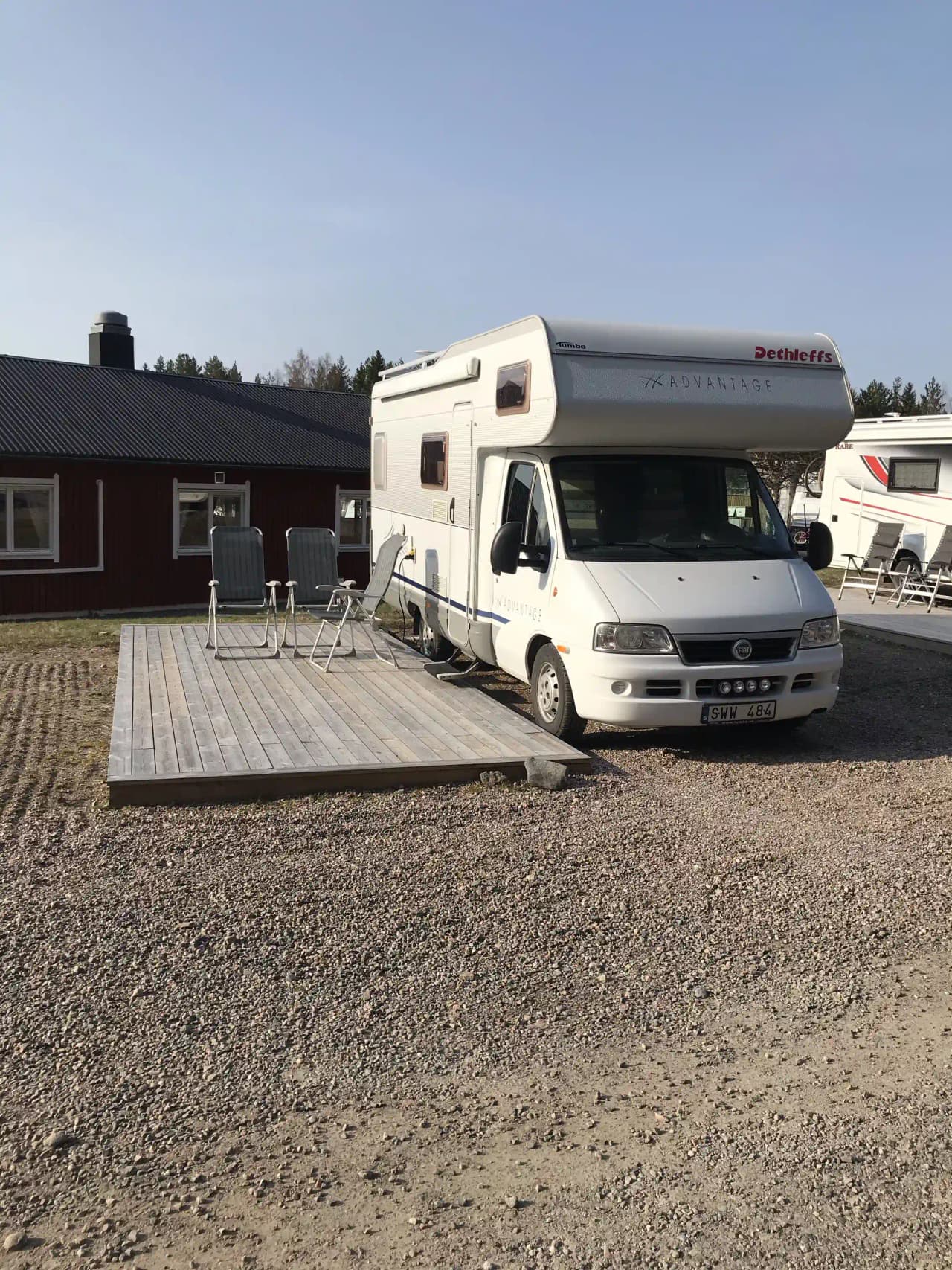 En rad med husvagnar och husbilar uppställda på en parkeringsplats vid Jogersö Eco Camping, omgivna av frodig grönska och klar himmel.
