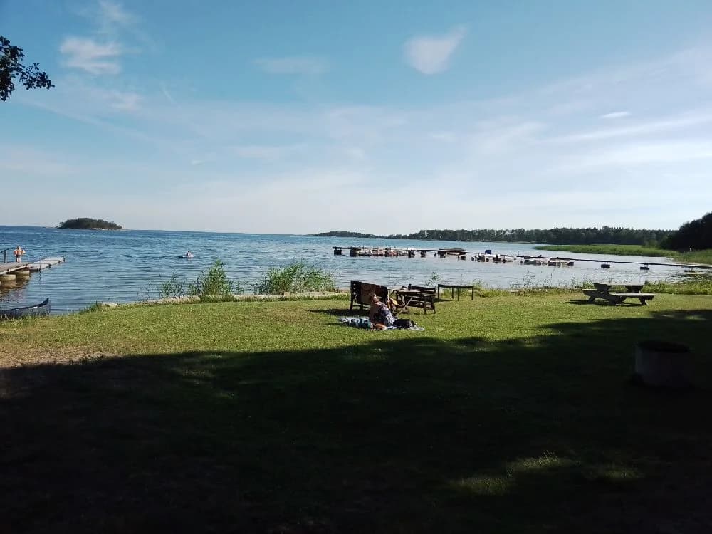 En somrig vy vid Jogersö Eco Camping med en sjö i förgrunden, omgiven av grönska och en sandig strandkant. Utomhusmöbler är placerade nära vattnet för rekreation och avkoppling.