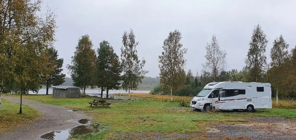 En husbil och en husvagn parkerade på grus vid Sikeå Havscamping, omgivna av gröna träd och omedelbar närhet till en våtmarksmiljö.