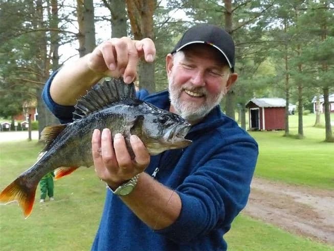 En person fiskar vid en sjö på Björkebo Camping, omgiven av natursköna omgivningar. En abborre syns i vattnet, vilket illustrerar det rika fiskelivet och de rekreationsmöjligheter som campingen erbjuder.