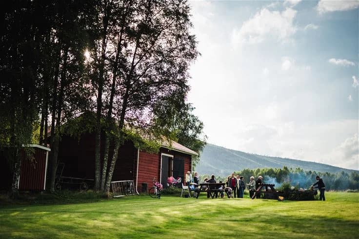 En pittoresk stuga på Björkebo Camping, omgiven av en grönskande gräsmatta och äng, med lantliga byggnader och en lada i bakgrunden.