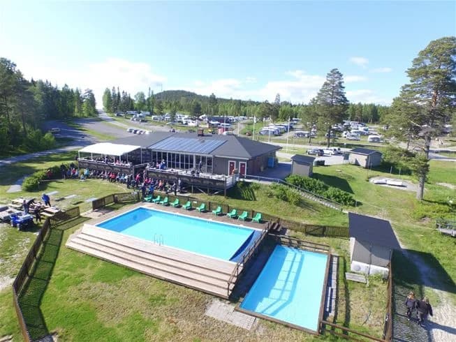 En solig dag vid Gullviks Havsbad Camping & Stugby, där en utomhuspool omgiven av solstolar och parasoller erbjuder avkoppling och nöje. Semesterfirare njuter av den avslappnade atmosfären i detta natursköna och miljövänliga semesterparadis.