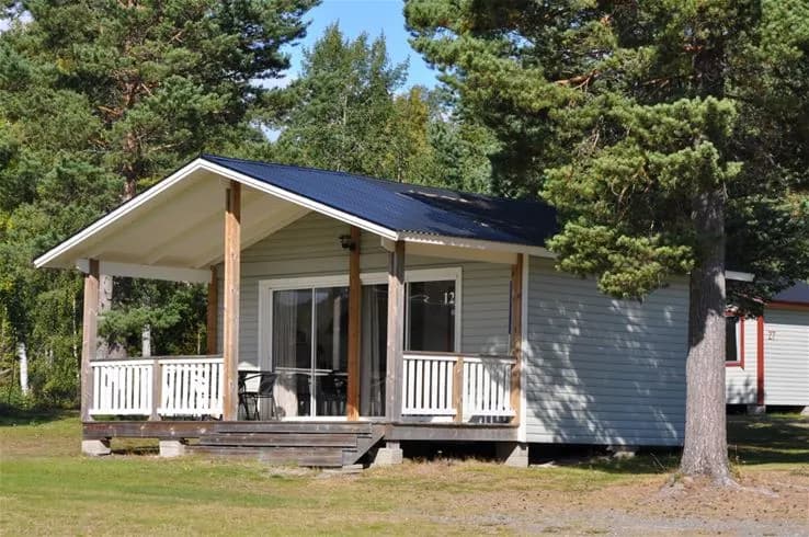 Bild av en stuga på Gullviks havsbad camping & stugby, med en veranda och trädörr. Stugan är omgiven av frodig växtlighet och träpaneler pryder dess utsida.