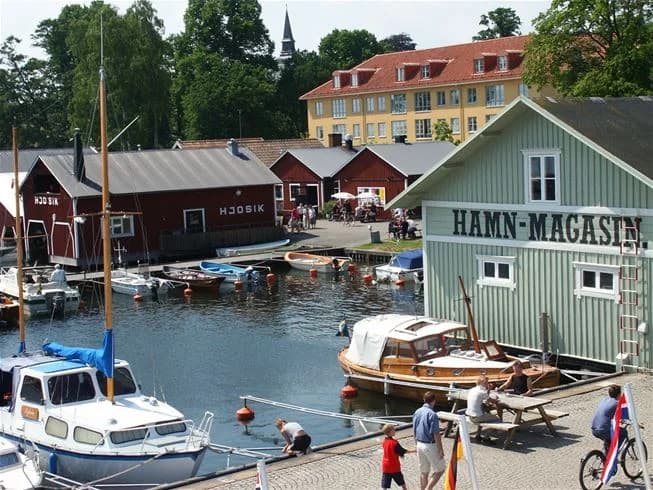 Bild av ett livligt marint område vid Breviks camping & ställplats, med flera motorbåtar som ligger förtöjda vid en brygga. Vattnet glittrar under en klar himmel, och området erbjuder både båtplatser och naturskön utsikt.