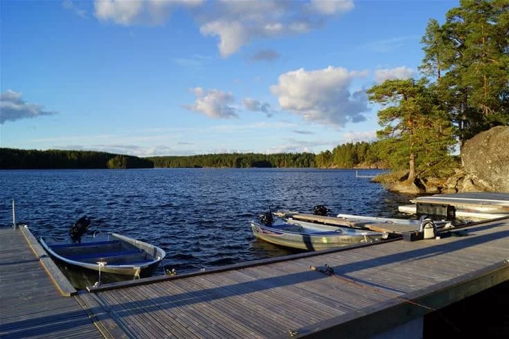 En rofylld sjö med glittrande vatten omgiven av lummiga träd. En liten brygga sträcker sig ut från stranden, perfekt för avkoppling och bad.