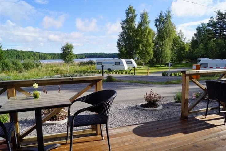 Alt-text: En utomhusmiljö vid Långsjön Stugor & Camping med bord och stolar i trä vid en sjö. Bakgrunden visar en lugn vattenspegel och omgivande grönska, vilket skapar en inbjudande och avkopplande atmosfär.