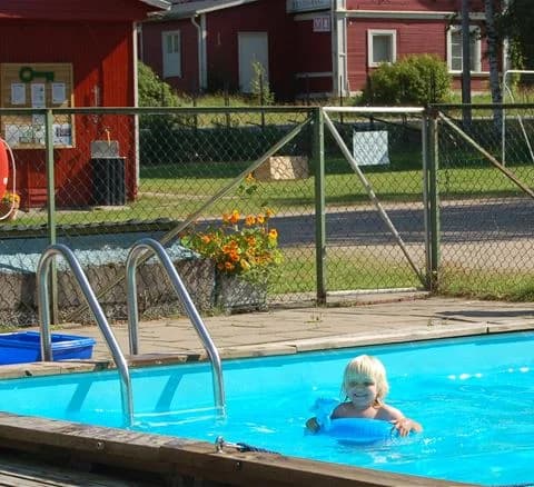 En solig campingbild med en pool där människor njuter av fritid och semester. Det finns solsängar runt poolen, och några besökare bär badkläder och leker i vattnet, vilket skapar en avslappnad och glad atmosfär.