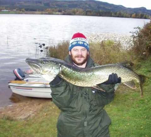 En fiskare håller upp en nyfångad gädda vid en sjö, omgiven av natur och campingmöjligheter.
