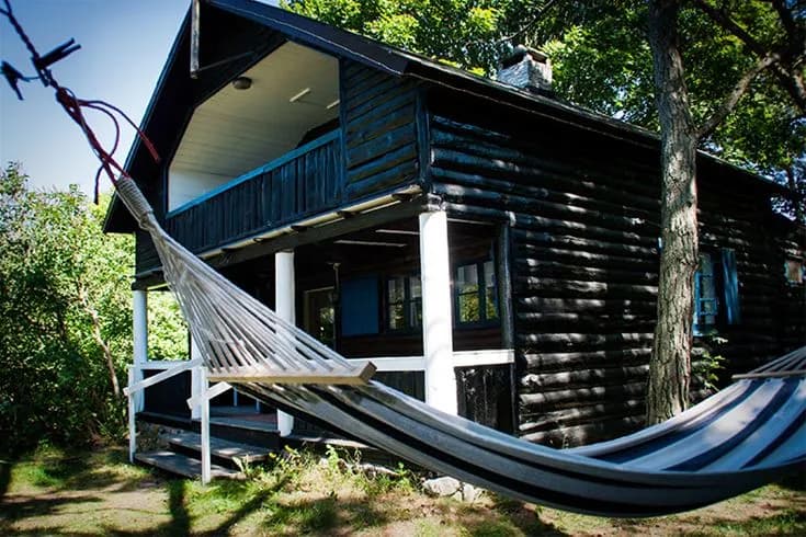 En mysig timmerstuga med veranda omgiven av grönska, belägen på Tofta camping. Träpaneler och skuggiga områden kompletterar den naturnära atmosfären.